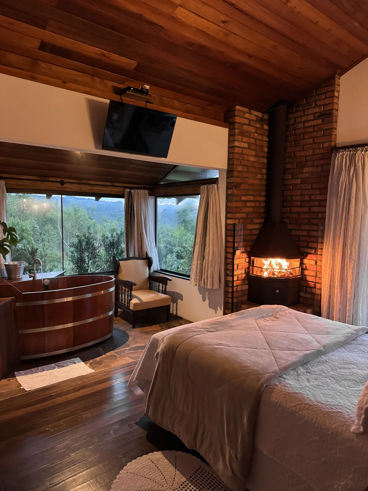A warm timber bedroom with a fireplace, soaking tub, and forest view